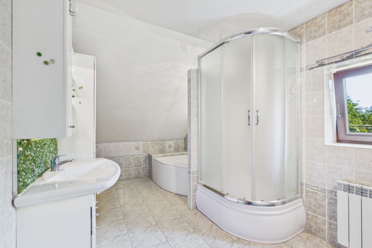 Bright bathroom with curved glass shower enclosure, white tub, and sink; tiled walls and floor with a green mosaic accent near the sink and a window to the right