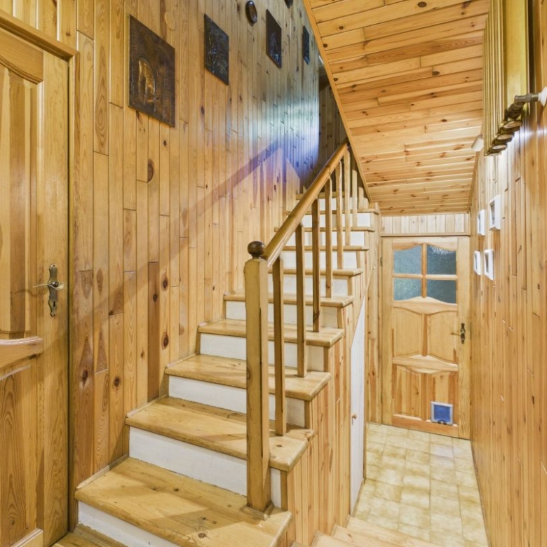 Wood-paneled hallway with a wooden staircase and rail leading up, warm natural tones throughout the walls and ceiling in a cozy cabin style hallway. It shows a door at the end and tiled floor along the corridor.