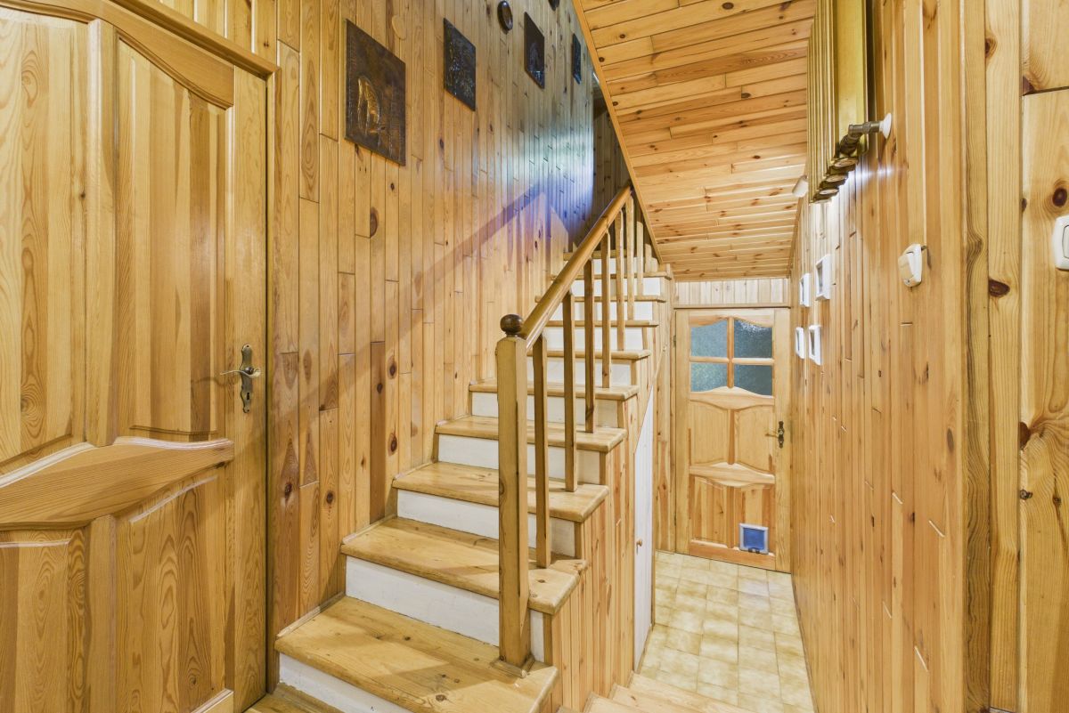 Wood-paneled hallway with a wooden staircase and rail leading up, warm natural tones throughout the walls and ceiling in a cozy cabin style hallway. It shows a door at the end and tiled floor along the corridor.