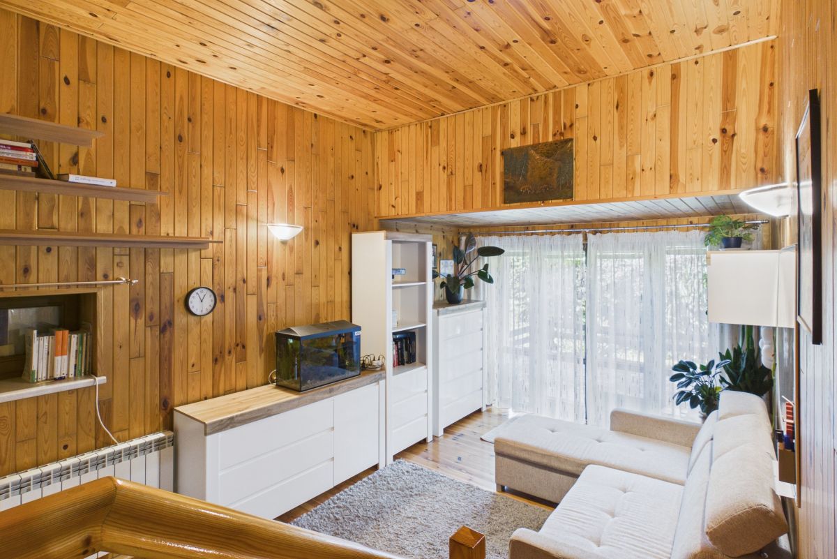 Cozy living room with wooden panel walls, white cabinets, and a beige sofa facing large glass doors with sheer curtains.