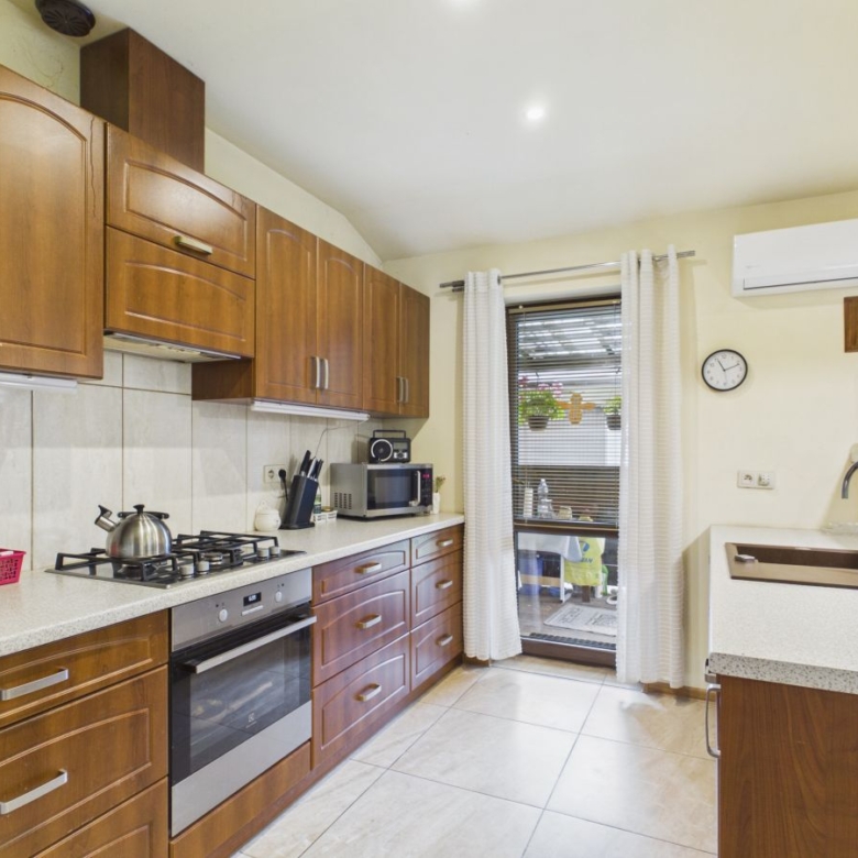 Kitchen with wooden cabinets, gas stove, oven, and kettle on a light counter; window with blinds and curtains at the back.