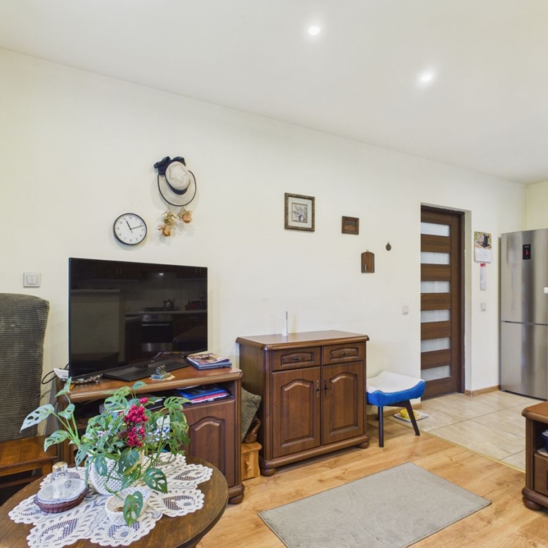 Cozy living room with TV on a wooden stand, round coffee table with a lace doily and flowers, and a kitchen with a stainless fridge in the background.