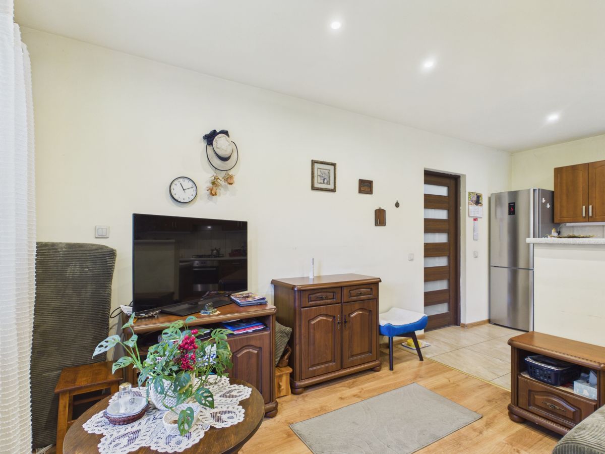 Cozy living room with TV on a wooden stand, round coffee table with a lace doily and flowers, and a kitchen with a stainless fridge in the background.