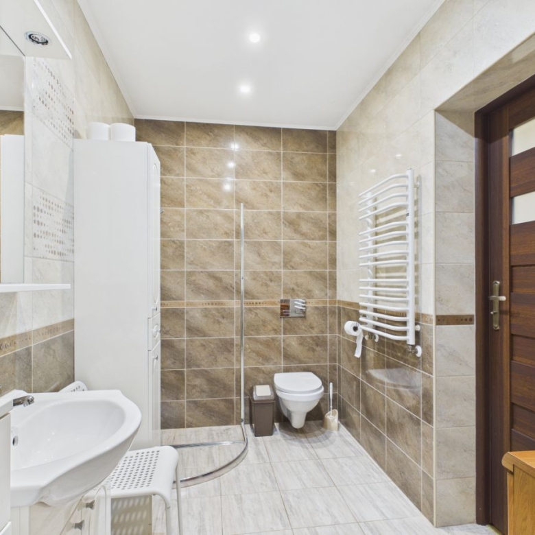 Modern bathroom with a corner curved shower, wall-mounted toilet, white sink, and tall white storage cabinet; a heated towel rack on the right wall and a wooden door to the room.