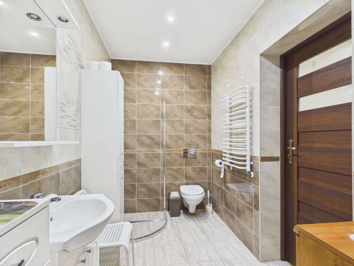 Modern bathroom with a corner curved shower, wall-mounted toilet, white sink, and tall white storage cabinet; a heated towel rack on the right wall and a wooden door to the room.