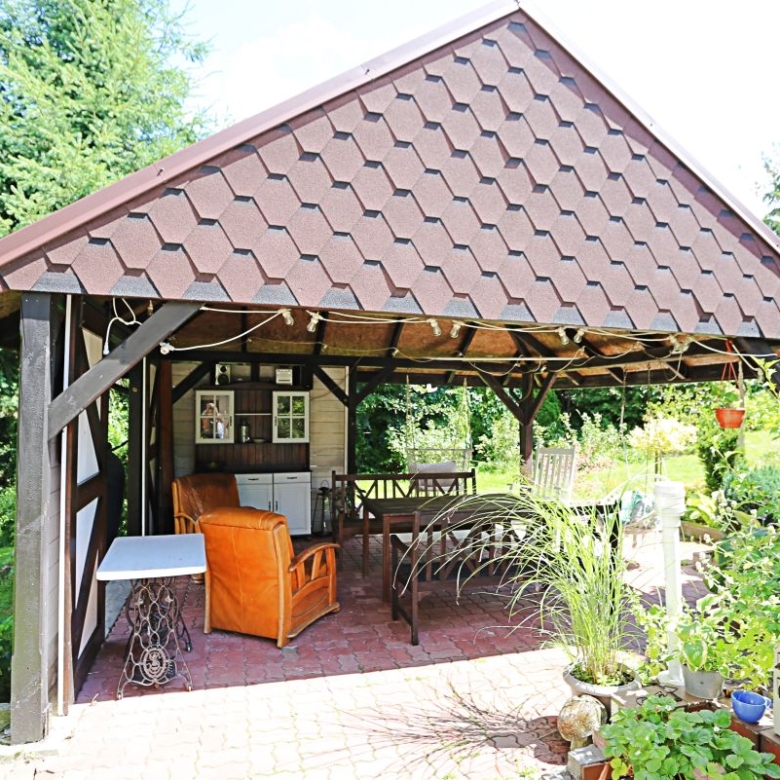 Wood gazebo with a brown tiled roof in a sunny garden, featuring outdoor seating and potted plants inside or nearby.
