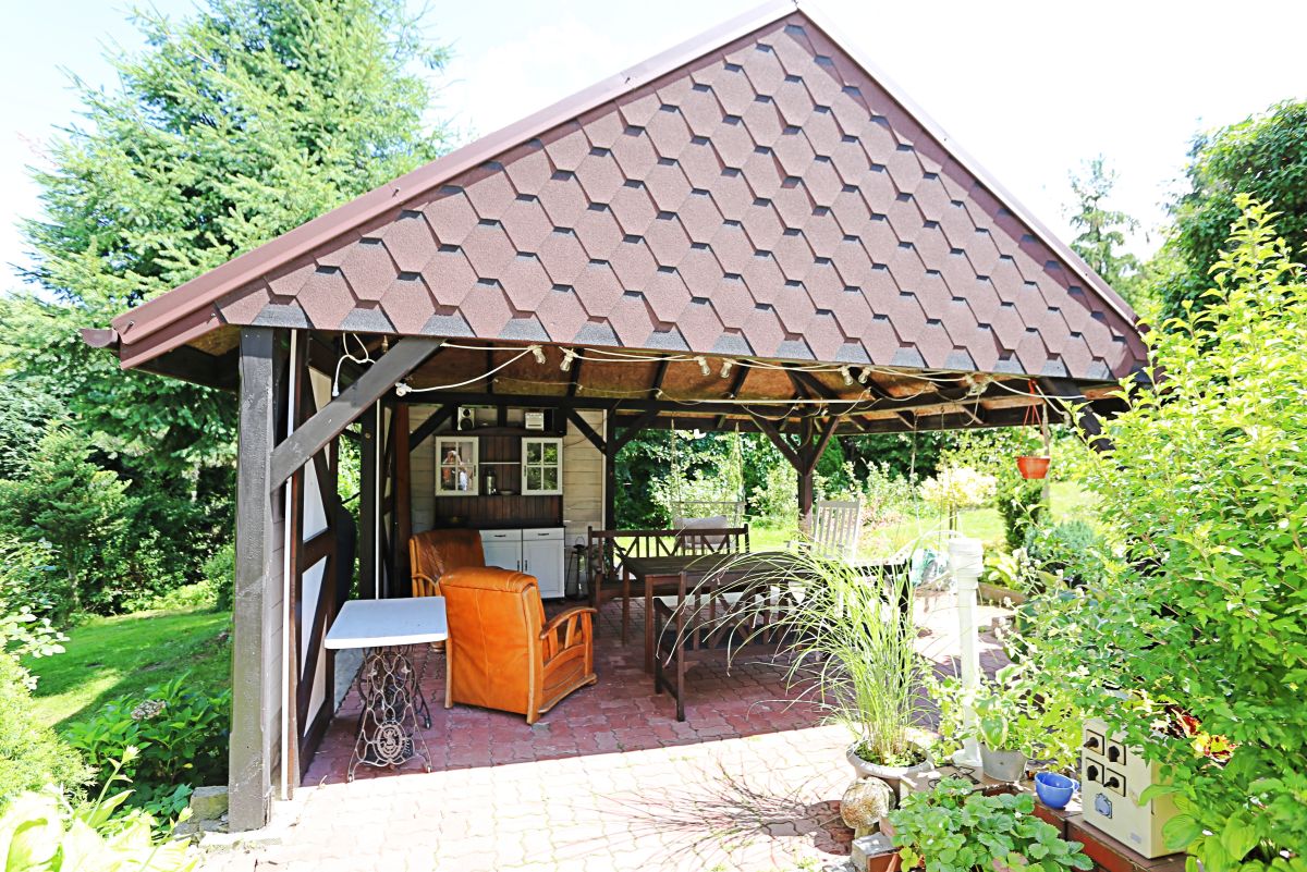 Wood gazebo with a brown tiled roof in a sunny garden, featuring outdoor seating and potted plants inside or nearby.