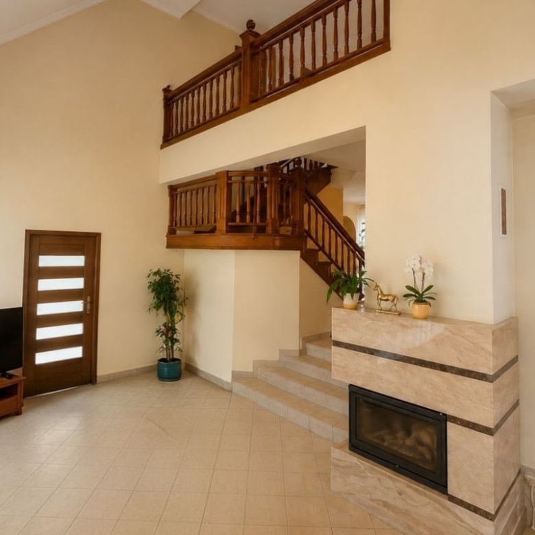 Bright living room with a two-story wood balcony, staircase, and a stone-framed fireplace; TV on a wooden stand near a door with horizontal glass panels.