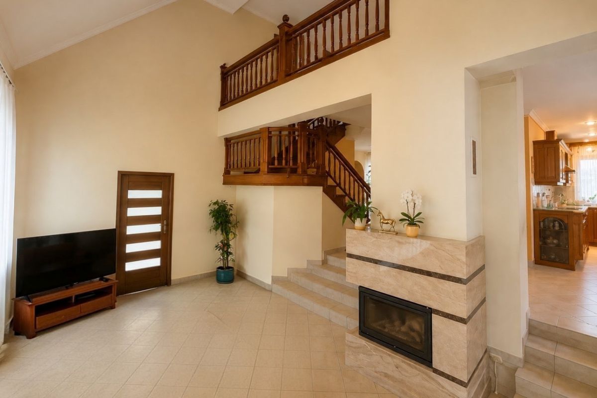 Bright living room with a two-story wood balcony, staircase, and a stone-framed fireplace; TV on a wooden stand near a door with horizontal glass panels.