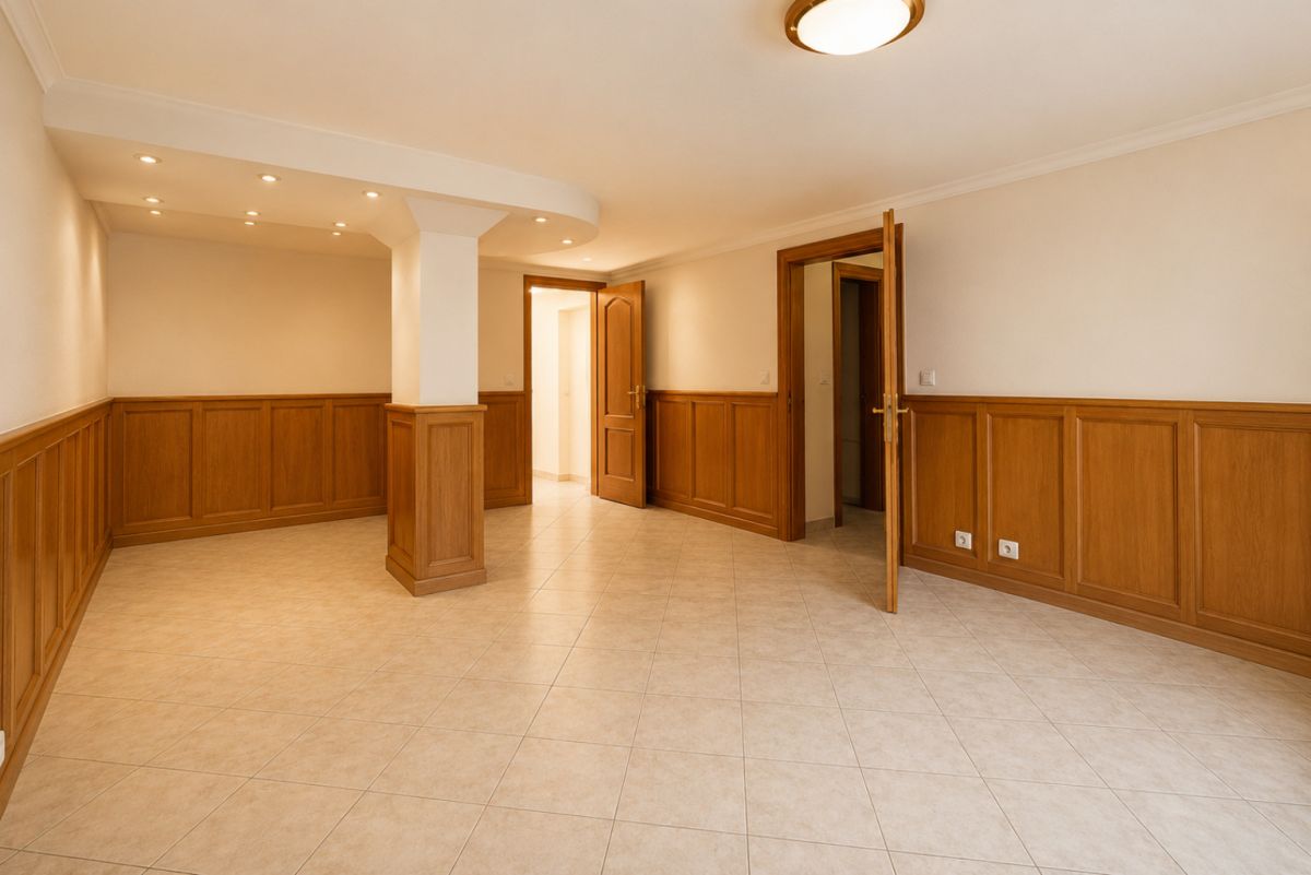 Empty, well-lit room with beige walls, wood paneling along the lower half, and a central column; two open doors and tiled floor.