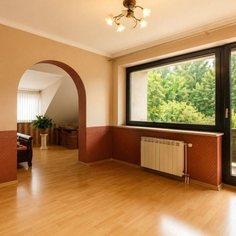 Sunlit living room with beige walls, red decorative wainscoting, and an arched doorway leading to a seating area; large window/door opens to a balcony with greenery outside.
