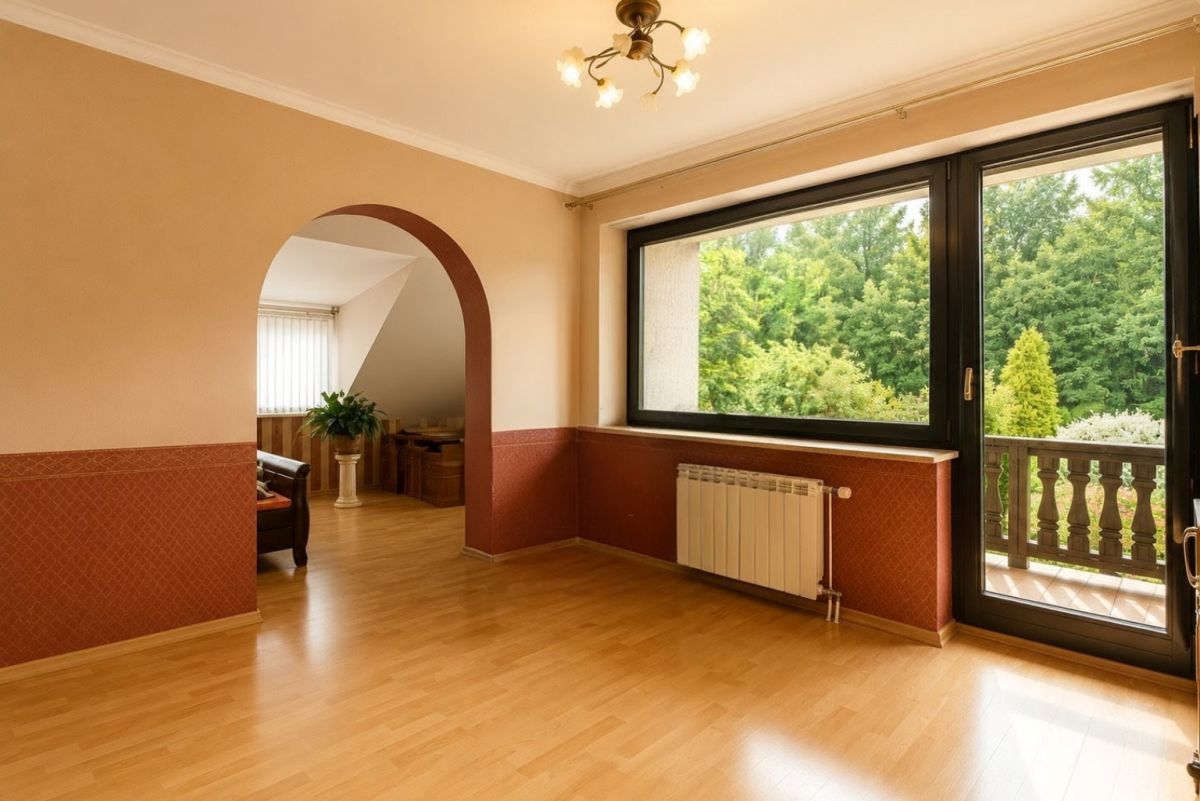 Sunlit living room with beige walls, red decorative wainscoting, and an arched doorway leading to a seating area; large window/door opens to a balcony with greenery outside.