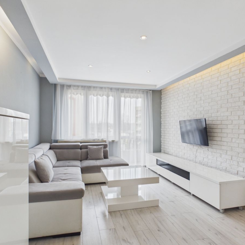 Modern living room with white brick wall, gray sectional sofa, and hanging TV on a white media console. Large windows with sheer curtains fill the space with light.