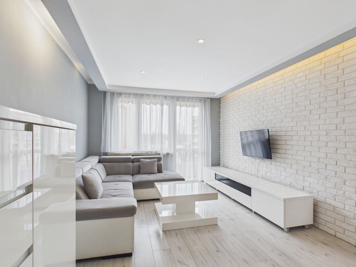 Modern living room with white brick wall, gray sectional sofa, and hanging TV on a white media console. Large windows with sheer curtains fill the space with light.