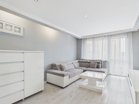 Bright minimalist living room with a light gray L-shaped sofa, white coffee table, and sheer-curtained windows letting in daylight.