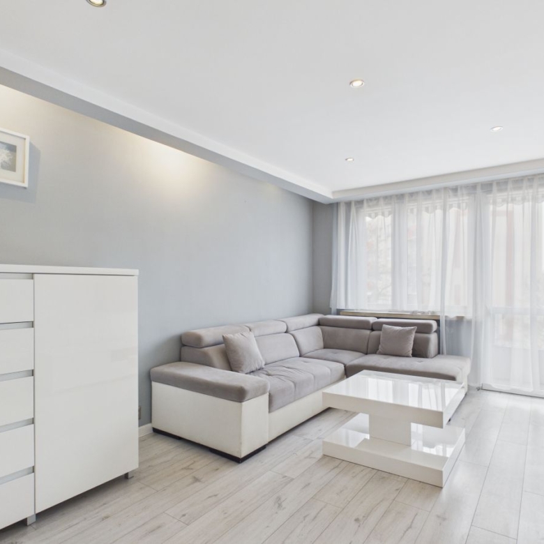 Bright minimalist living room with a light gray L-shaped sofa, white coffee table, and sheer-curtained windows letting in daylight.