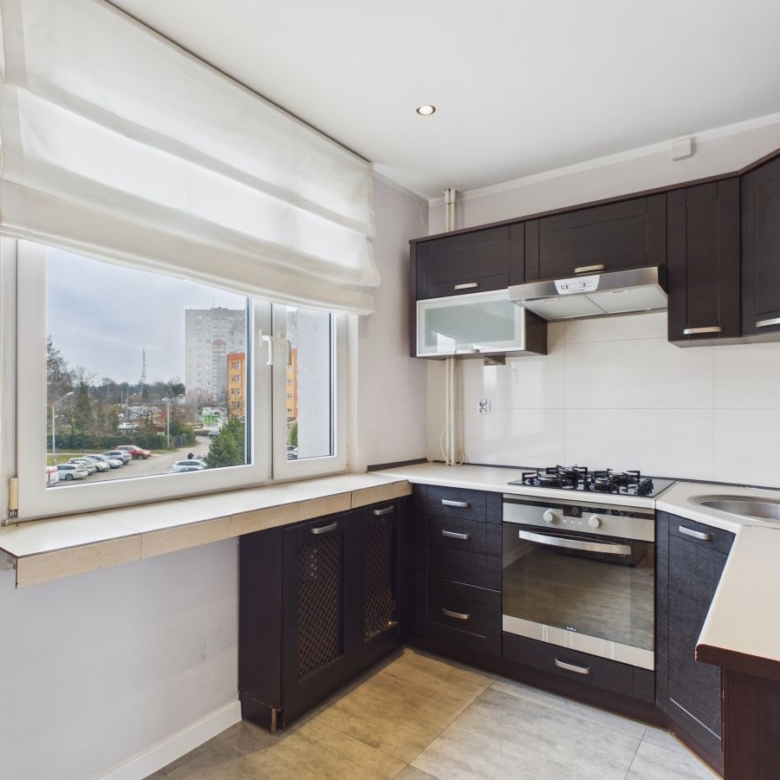 Modern L-shaped kitchen with dark cabinetry, white countertops, gas stove and built-in oven, stainless sink, and large window with a white shade overlooking apartments outside.