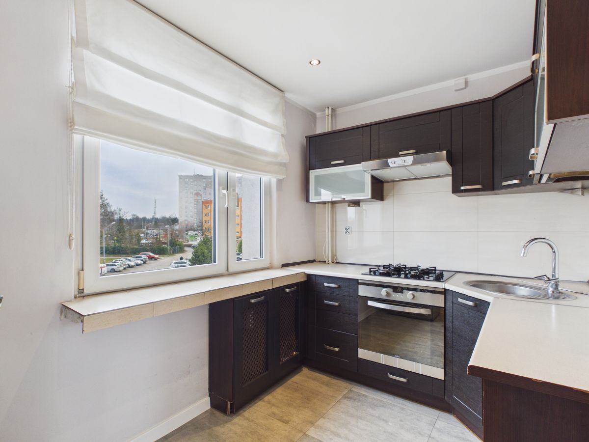 Modern L-shaped kitchen with dark cabinetry, white countertops, gas stove and built-in oven, stainless sink, and large window with a white shade overlooking apartments outside.