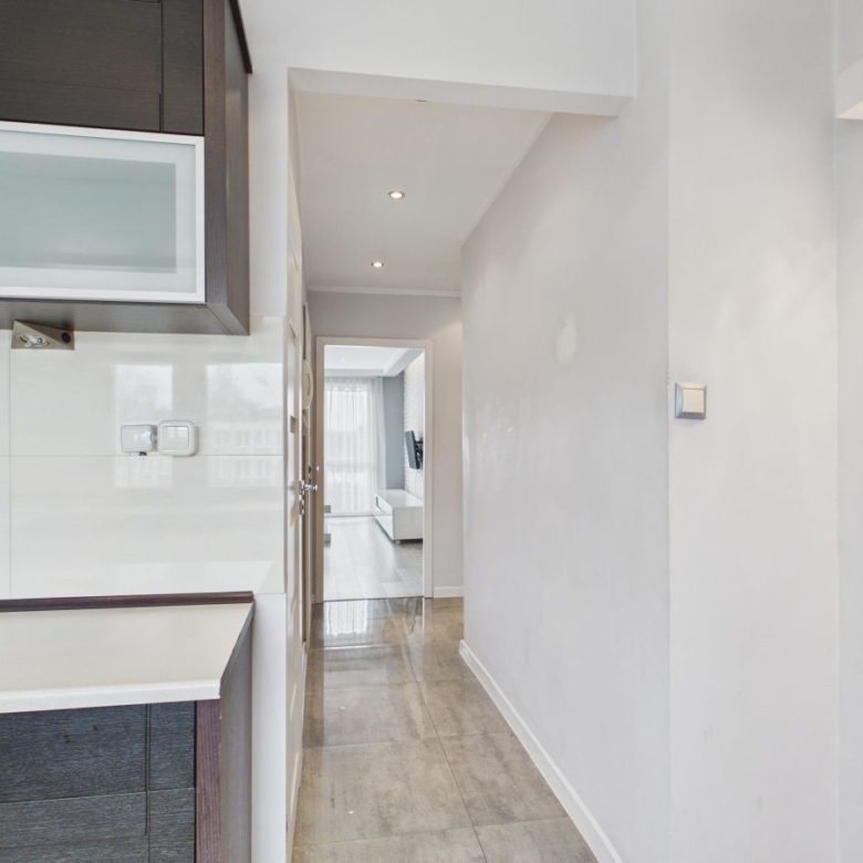 Modern kitchen with dark wood cabinets on the left, white backsplash, and a bright living area at the end of the hallway.