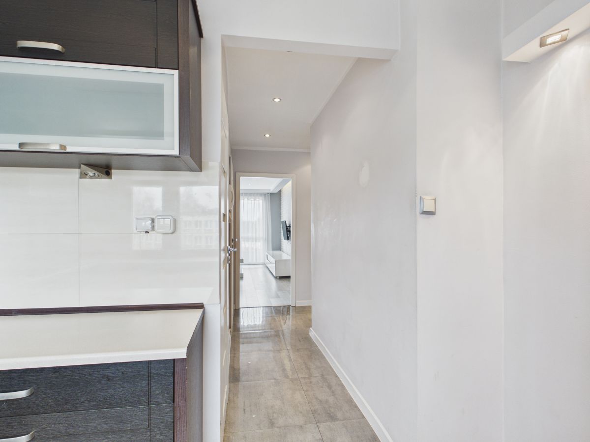Modern kitchen with dark wood cabinets on the left, white backsplash, and a bright living area at the end of the hallway.
