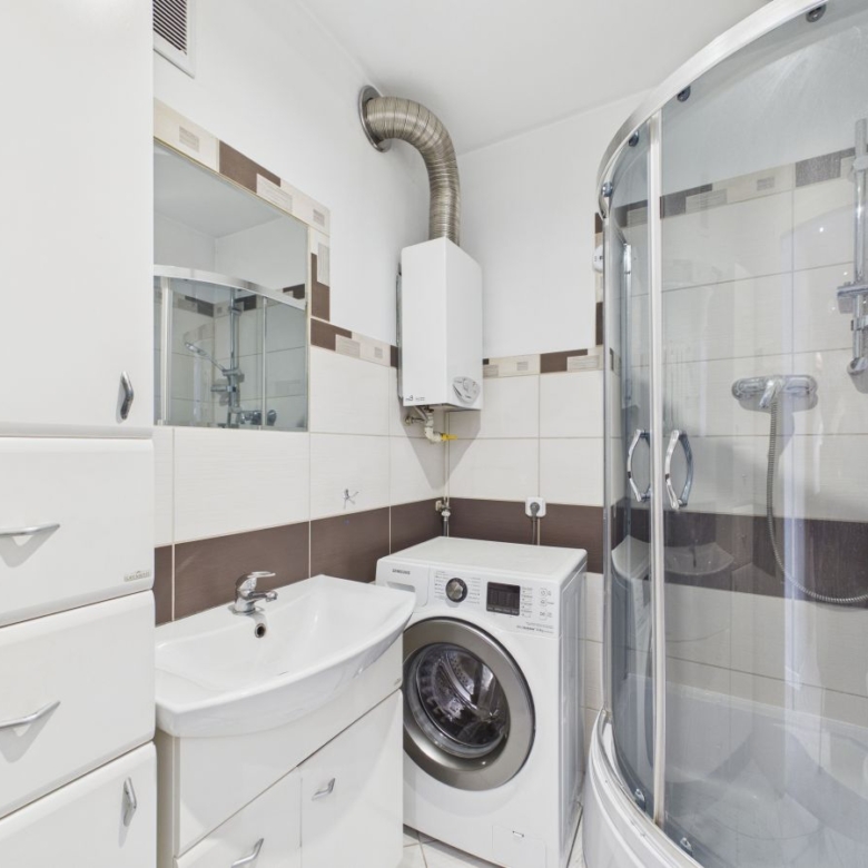 Small modern bathroom with curved glass shower enclosure, washing machine, white sink, and tall storage drawers; white tiles with brown accents.