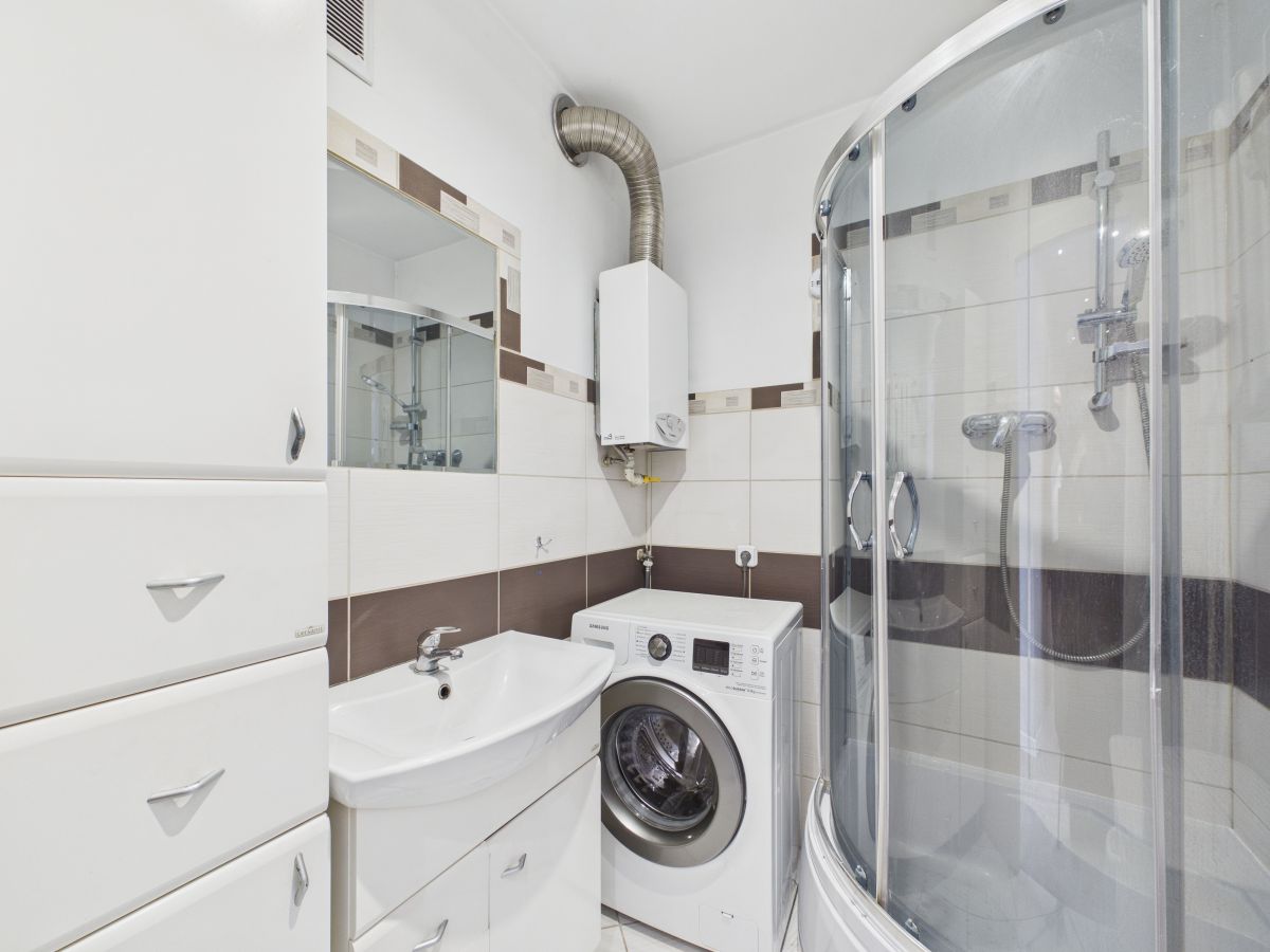 Small modern bathroom with curved glass shower enclosure, washing machine, white sink, and tall storage drawers; white tiles with brown accents.
