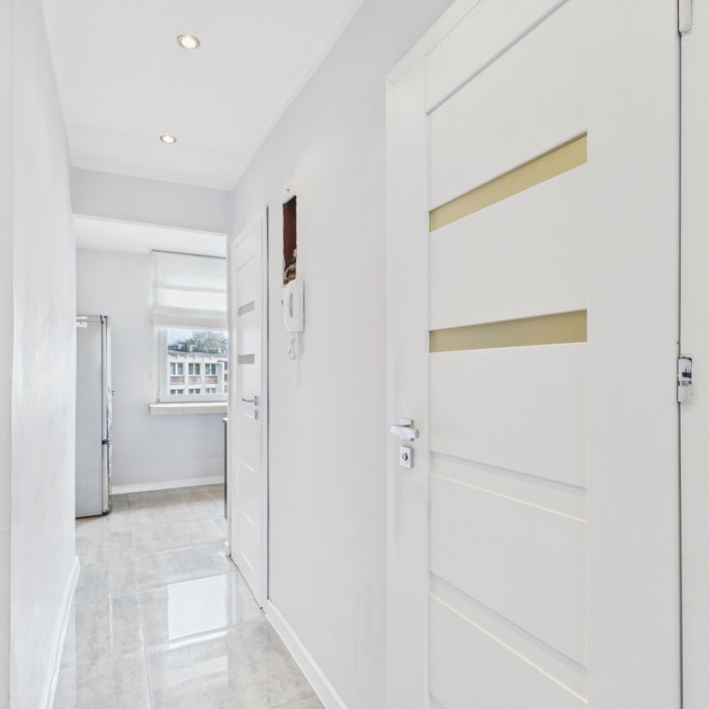 Bright white hallway with glossy tiled floor leading to a kitchen; a tall fridge visible by a window at the end.