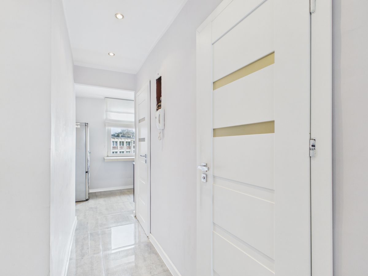 Bright white hallway with glossy tiled floor leading to a kitchen; a tall fridge visible by a window at the end.