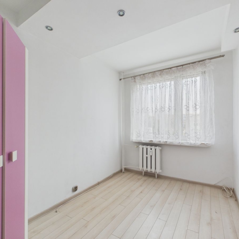 Small empty room with light wood floor, white walls, and a pink closet on the left; lace-curtained window and radiator beneath it in the corner.