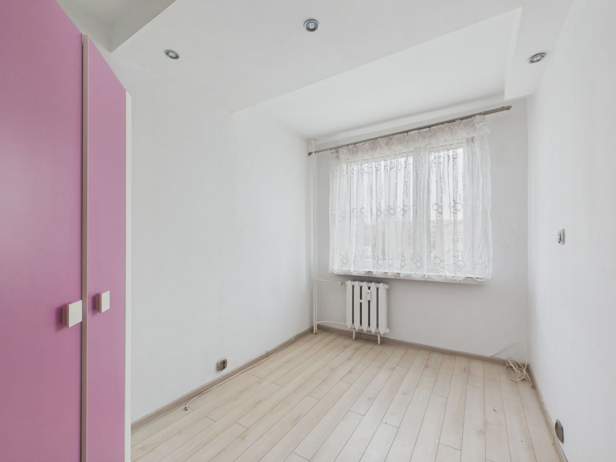 Small empty room with light wood floor, white walls, and a pink closet on the left; lace-curtained window and radiator beneath it in the corner.