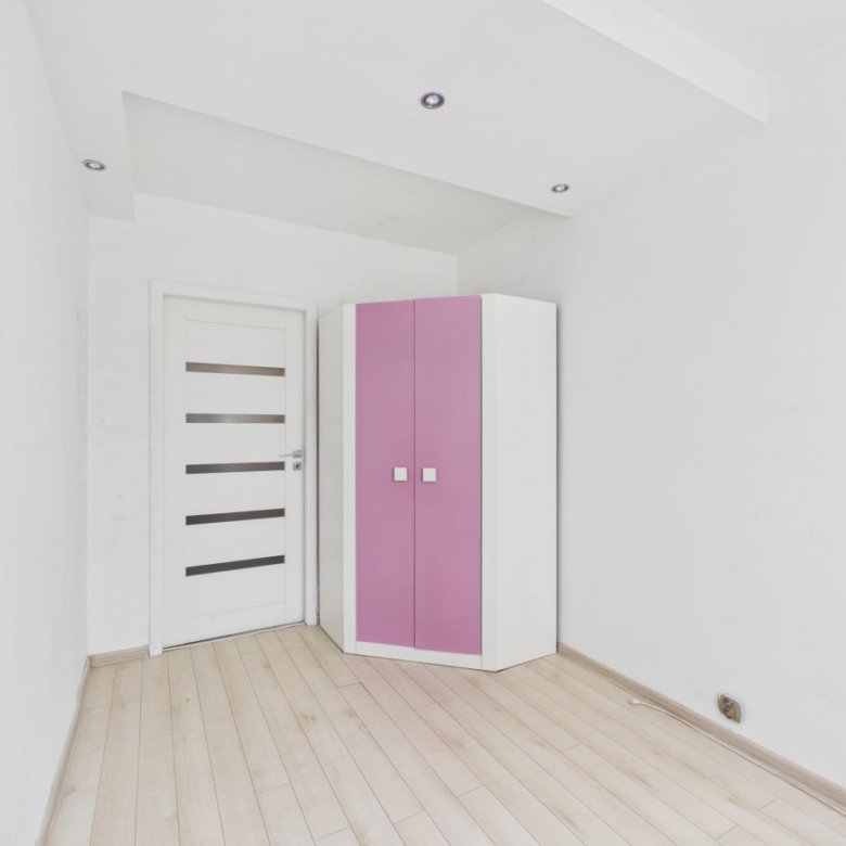 A minimal white room with a white door featuring horizontal glass panels and a pink-and-white corner wardrobe near the corner.