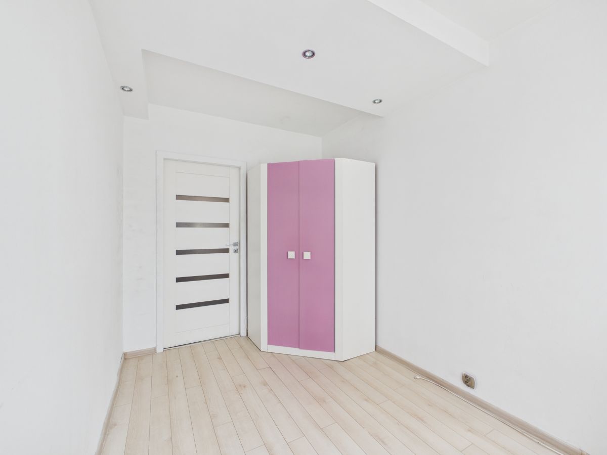 A minimal white room with a white door featuring horizontal glass panels and a pink-and-white corner wardrobe near the corner.