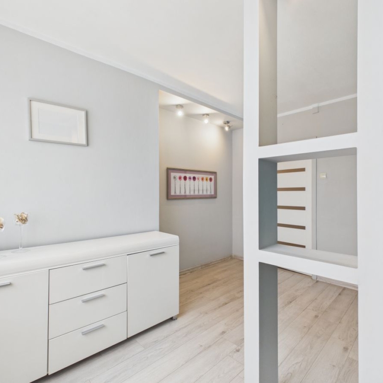Bright minimalist hallway with a white sideboard along the left wall, decorative vases, and light-toned wooden flooring viewed through a white grid divider in the foreground.