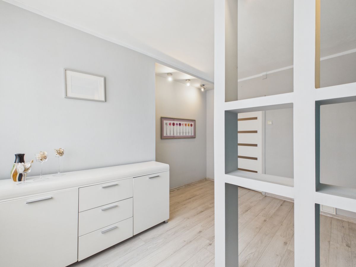Bright minimalist hallway with a white sideboard along the left wall, decorative vases, and light-toned wooden flooring viewed through a white grid divider in the foreground.