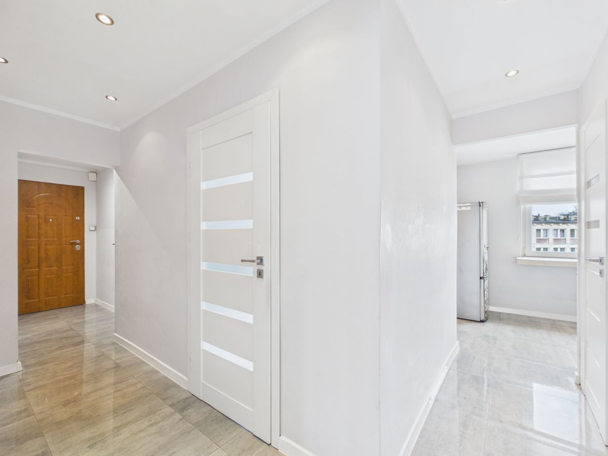 Bright hallway in a modern apartment with white walls, a wooden front door on the left, and a white interior door with horizontal frosted-glass panels in the center.
