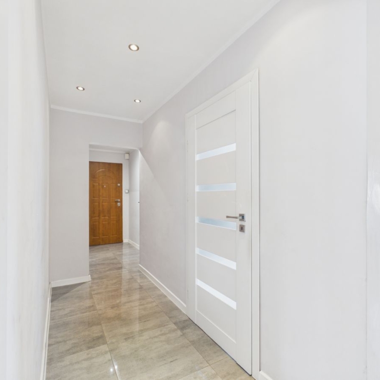 Bright hallway with gray-tiled floor, white walls, and a white door with frosted horizontal panels on the right; wooden door at the far end.