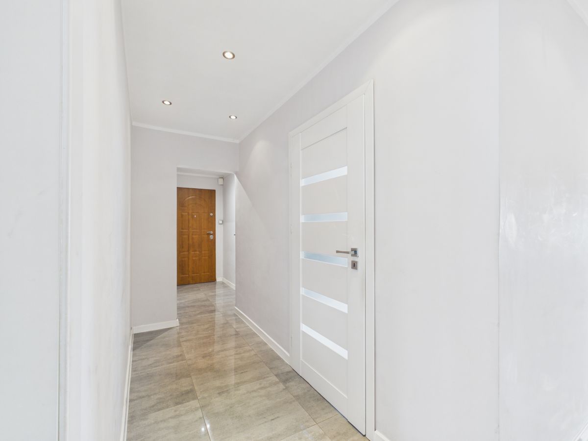 Bright hallway with gray-tiled floor, white walls, and a white door with frosted horizontal panels on the right; wooden door at the far end.