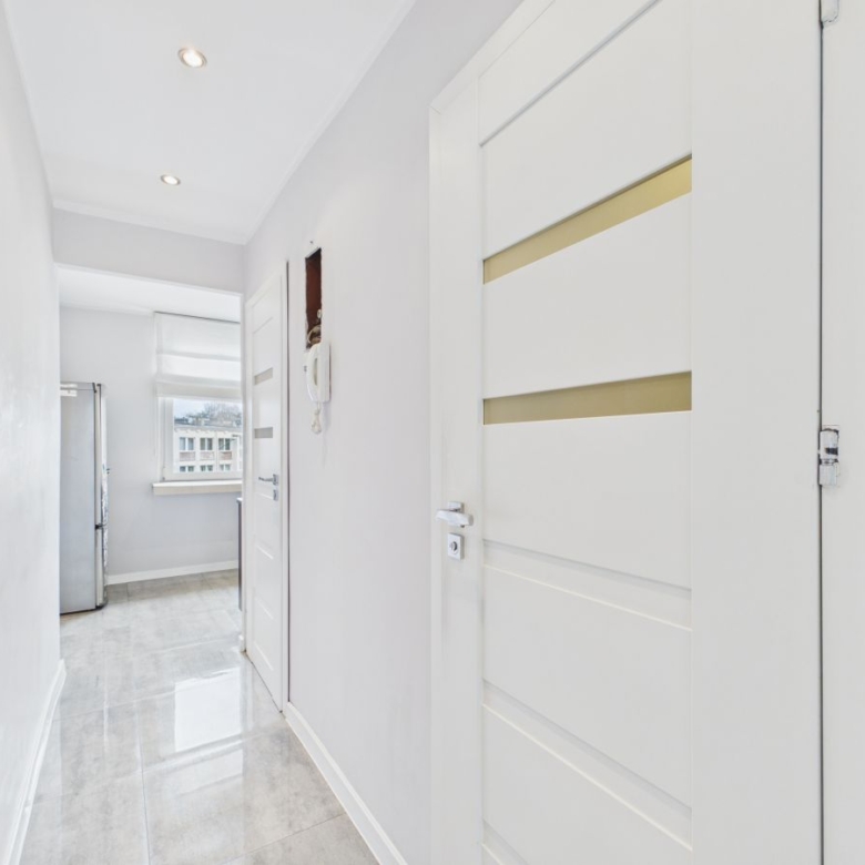 Bright white hallway with glossy tile floor, white interior doors, and a silver fridge visible near a window at the end of the hall failing to clutter the space.