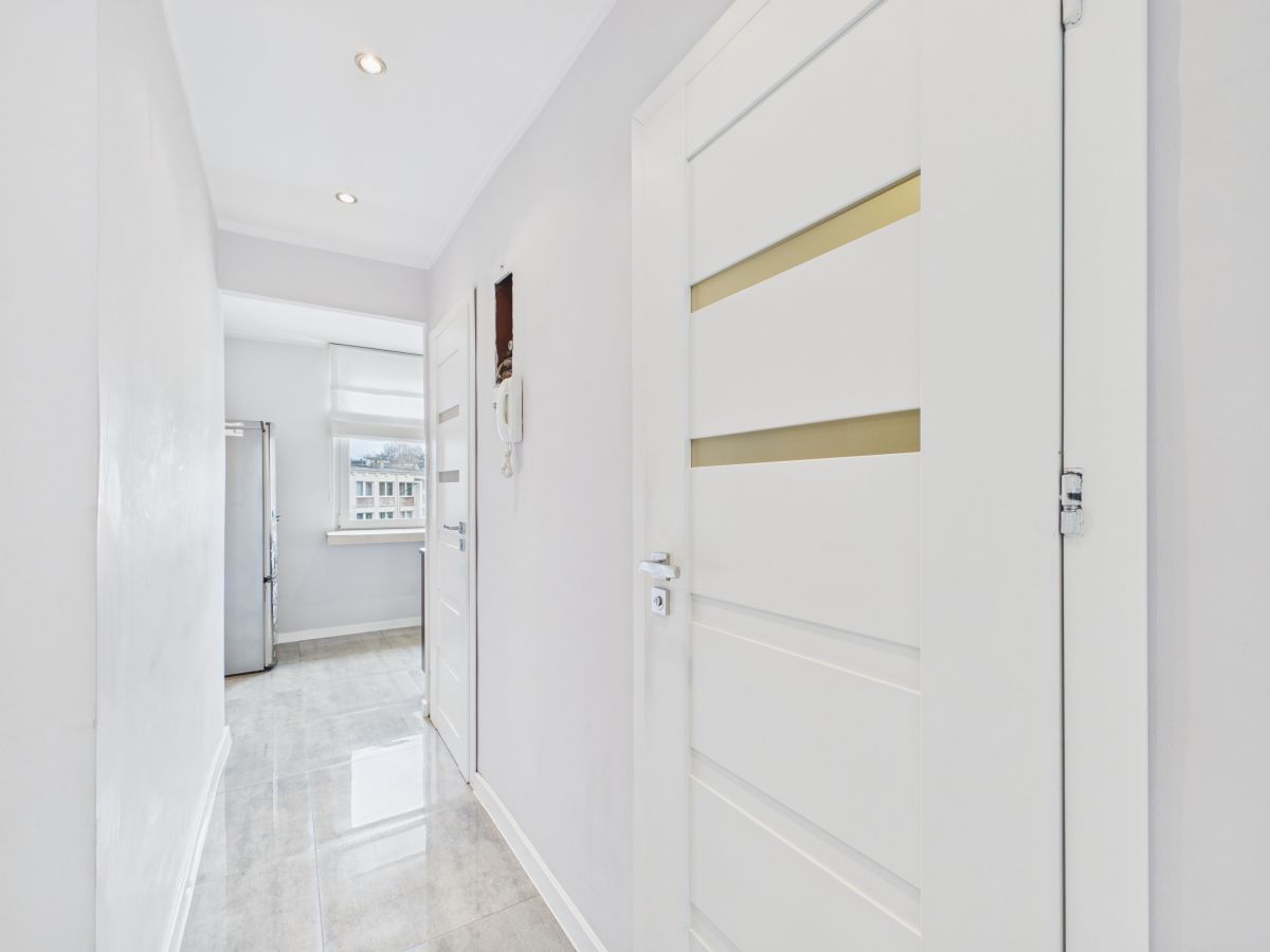 Bright white hallway with glossy tile floor, white interior doors, and a silver fridge visible near a window at the end of the hall failing to clutter the space.