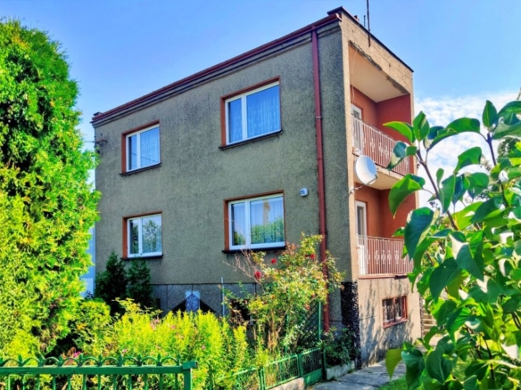 Two-story apartment building with beige-green facade, brown-framed windows, and a balcony on the right, surrounded by greenery.