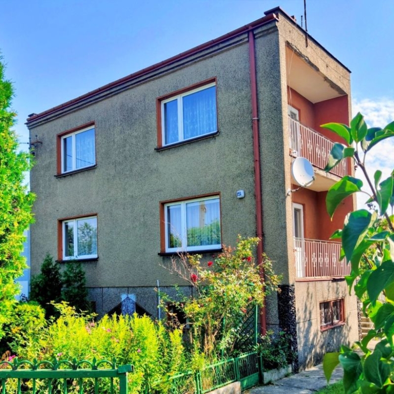Two-story apartment building with beige-green facade, brown-framed windows, and a balcony on the right, surrounded by greenery.