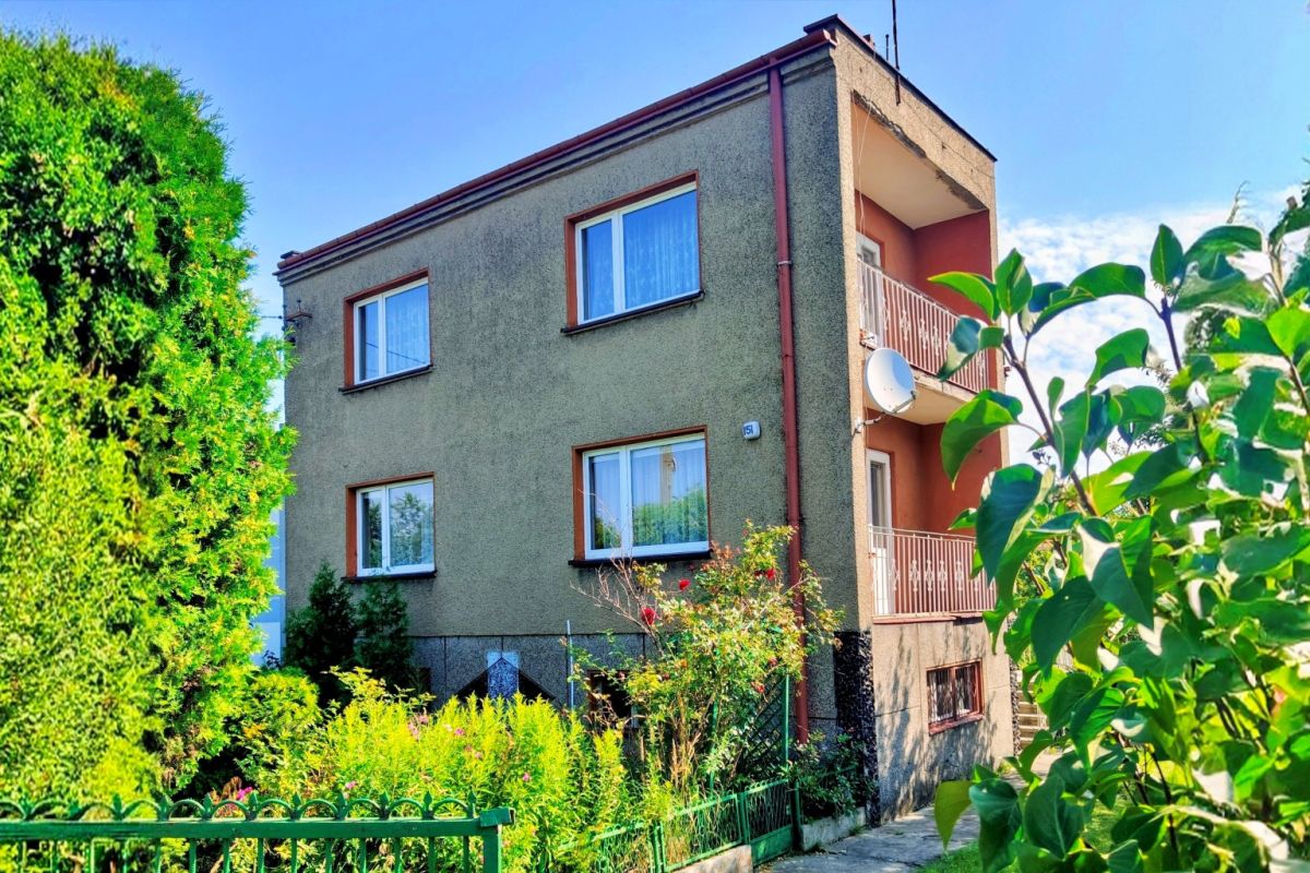 111832223.jpg Two-story apartment building with beige-green facade, brown-framed windows, and a balcony on the right, surrounded by greenery.
