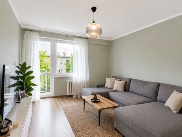 Bright living room with a gray sectional sofa, wooden coffee table, and sheer white curtains by a large window overlooking trees.