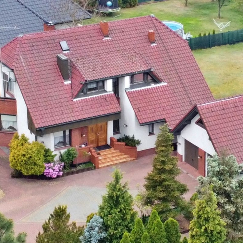 Aerial view of a suburban house with a red tiled roof, brick accents, and a manicured front yard surrounded by trees.