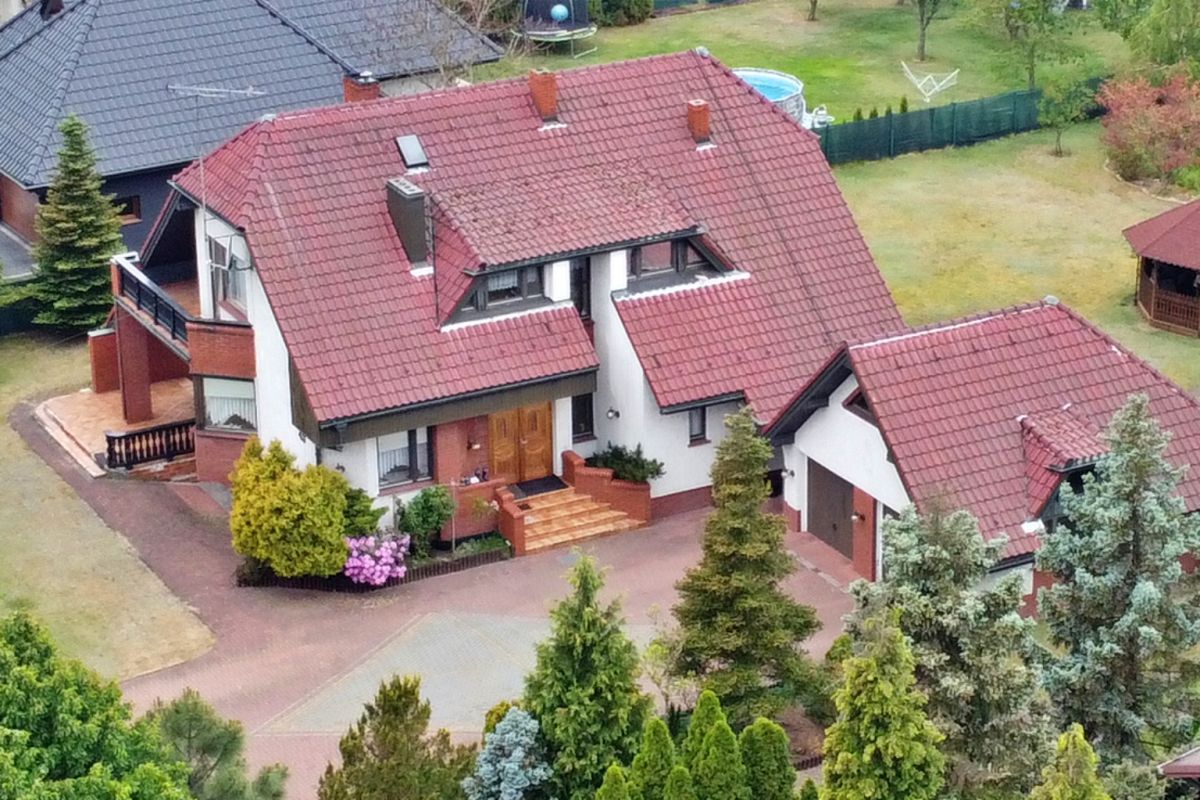 Aerial view of a suburban house with a red tiled roof, brick accents, and a manicured front yard surrounded by trees.