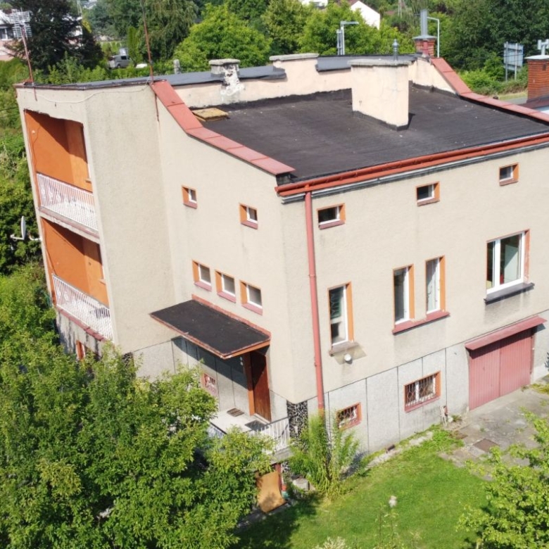 Aerial view of a beige, three-story apartment building with orange balconies, red gutters, and a flat black roof, surrounded by trees and greenery.