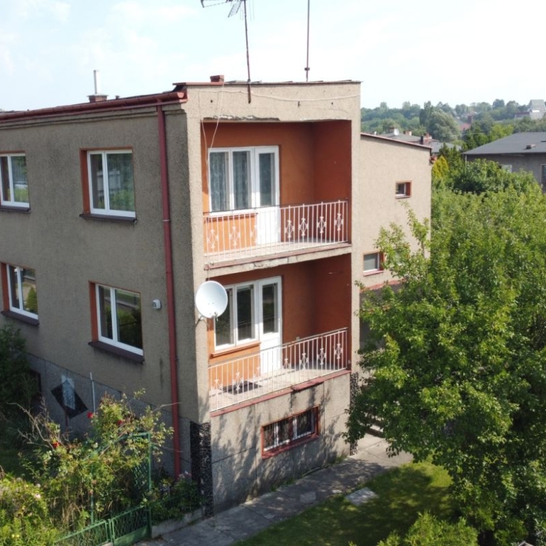 Three-story beige apartment building with two balconies, white railings, and a satellite dish in a tree-filled suburban setting.