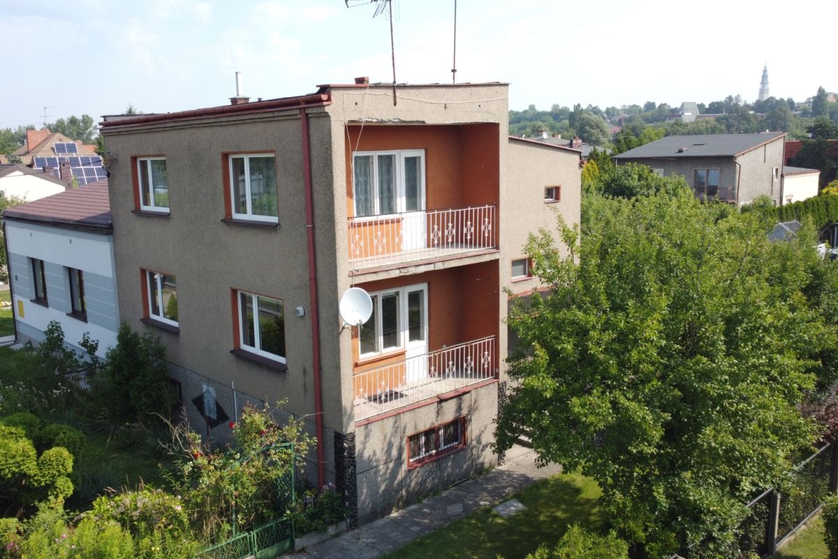 Three-story beige apartment building with two balconies, white railings, and a satellite dish in a tree-filled suburban setting.