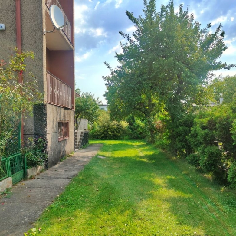 Apartment building on the left with a grassy lawn, a concrete path, and a satellite dish on a balcony, under a blue sky with trees along a hedge on the right.