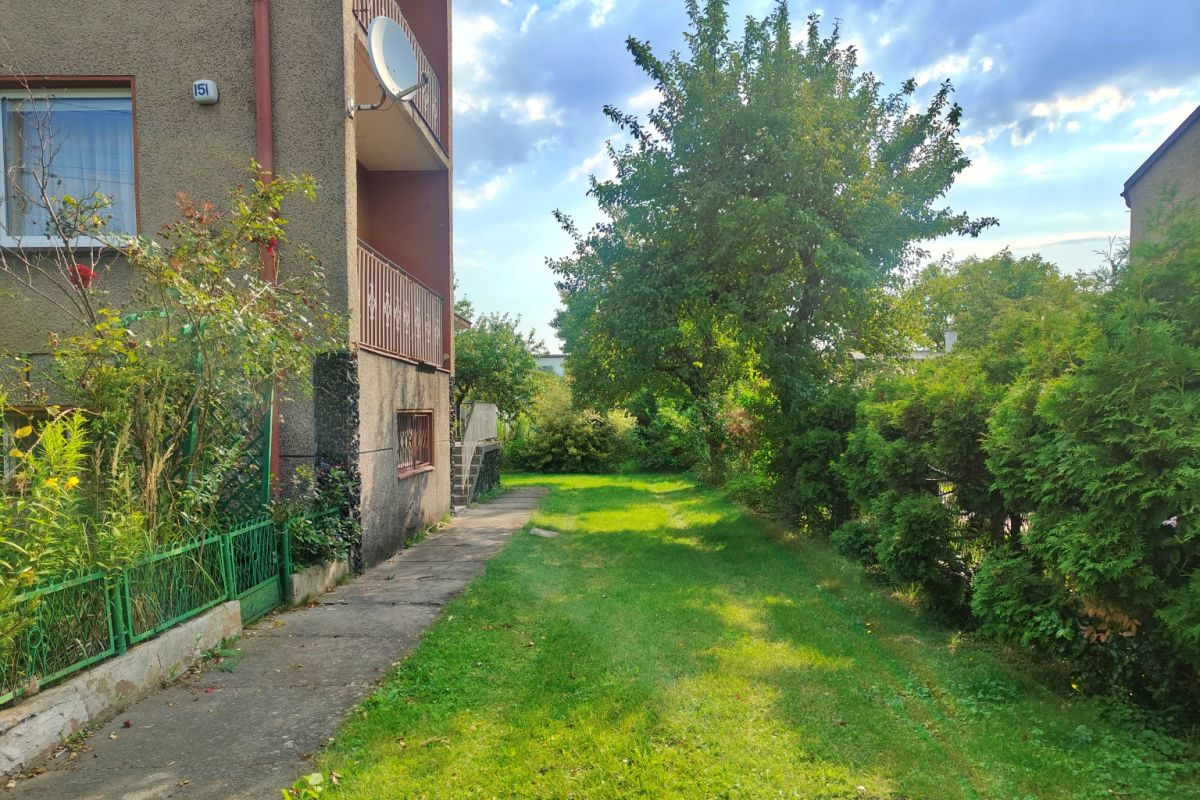 Apartment building on the left with a grassy lawn, a concrete path, and a satellite dish on a balcony, under a blue sky with trees along a hedge on the right.